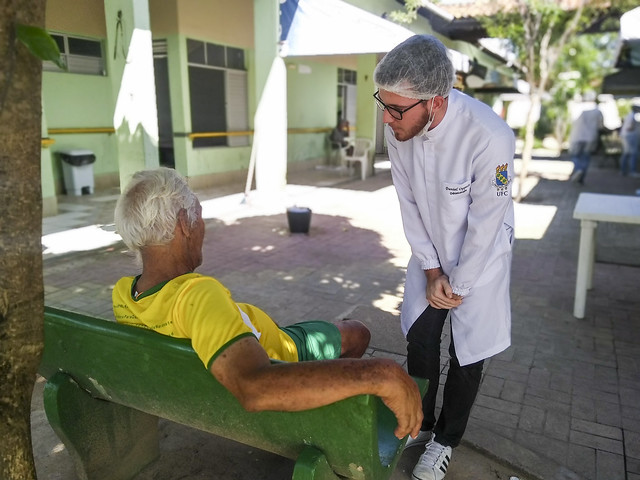 Ao todo, serão ofertadas até 645 bolsas, no valor de R$ 700,00 mensais, para o período de abril a dezembro de 2027 (Foto: Luana Oliveira/UFC) Imagem: um senhor de camisa amarela está sentado em um banco de costas para a foto. Em pé, um jovem de jaleco brando e touca conversa com ele (Foto: Luana Oliveira/UFC)