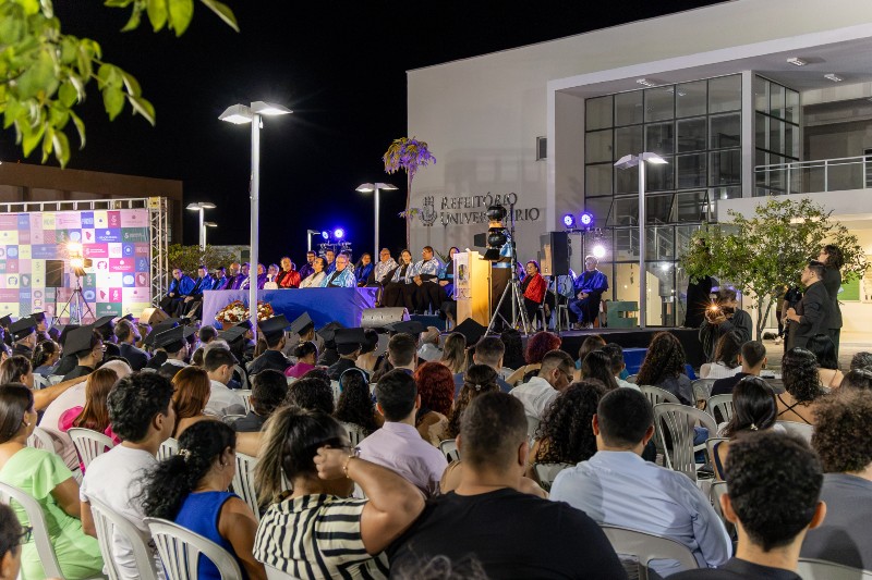 Cerimônia de Sobral foi realizada no Campus Mucambinho (Foto: Guilherme Silva/UFC) Imagem: Colação de grau da UFC em Sobral, realizada à noite. Formandos acompanham a cerimônia diante do palco, onde professores e autoridades acadêmicas participam da celebração.