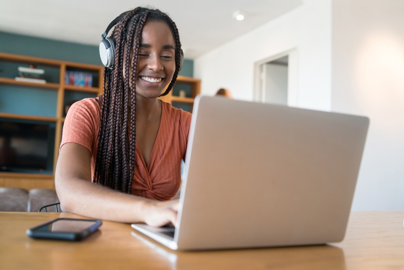 O curso é aberto a qualquer estudante estrangeiro interessado em obter certificação no Nível A1 do curso de Português (Imagem: Magnific) Imagem: Uma mulher negra, sorridente, usa fones de ouvido enquanto trabalha em um notebook sobre uma mesa de madeira. Ao fundo, há uma estante com livros e objetos decorativos em um ambiente residencial iluminado