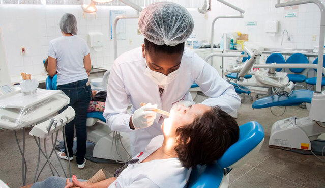 Substância da casca da castanha de caju pode ser usada para produzir como materiais resinas, adesivos e dessensibilizantes dentinários (Foto: Ribamar Neto/UFC) Imagem: Profissional de odontologia, usando máscara, luvas e touca, realiza exame bucal em uma paciente reclinada em uma cadeira odontológica. Ao fundo, há outros consultórios e pessoas em atendimento em uma clínica odontológica.
