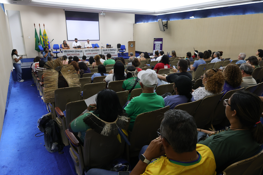 O evento integra a programação oficial da Missão Sobre a Instalação de Data Centers no Estado do Ceará, promovida pelo CNDH e pelo CEDDH. (Foto: Victor Braga/UFC) Imagem: auditório durante um seminário, com dezenas de pessoas sentadas acompanhando a programação. À frente, três participantes compõem a mesa de abertura, posicionada em um palco com telão de projeção ao fundo. Em frente à mesa, há faixas estendidas, e, ao lado esquerdo do palco, uma pessoa faz uso da palavra em um púlpito. Ao fundo do auditório, estão as bandeiras do Brasil, do Ceará e da Universidade Federal do Ceará (UFC). A plateia é composta por pessoas de diferentes perfis, algumas utilizando vestimentas e adereços de povos indígenas e movimentos sociais.