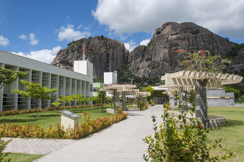 Imagem: área externa do campus de Quixadá, com céu azul e grandes formações rochosas ao fundo, parte da fachada de um prédio à esquerda e área da convivência com bancos à direita, tudo cercado por muitas plantas e grama