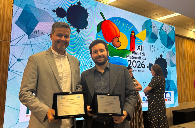 Professores Yuri Lima e Mauricio Poletti durante a cerimônia de entrega do Prêmio SBM 2026 (Foto: acervo pessoal) Foto mostra dois professores segurando certificados de premiação, em frente a um banner onde se pode ler: XII Bienal da Matemática (foto: Acervo pessoal)