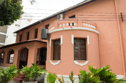 A modalidade de ensino será presencial (100%), e as aulas acontecerão exclusivamente às segundas e quartas-feiras, das 18h às 20h, na sede da Casa de Cultura Francesa (Foto: Jr. Panela/UFC Informa) Imagem: Fotografia da fachada da Casa de Cultura Francesa da Universidade Federal do Ceará. O prédio, de arquitetura histórica, é pintado em tom salmão e possui janelas de madeira, varanda semicircular com balaustrada branca e telhado de cerâmica. À esquerda, há uma varanda coberta sustentada por arcos, que dá acesso à entrada do edifício. Na parte frontal, jardins com plantas ornamentais verdes contornam a construção. Ao fundo, aparecem árvores, edificações vizinhas e fios da rede elétrica.