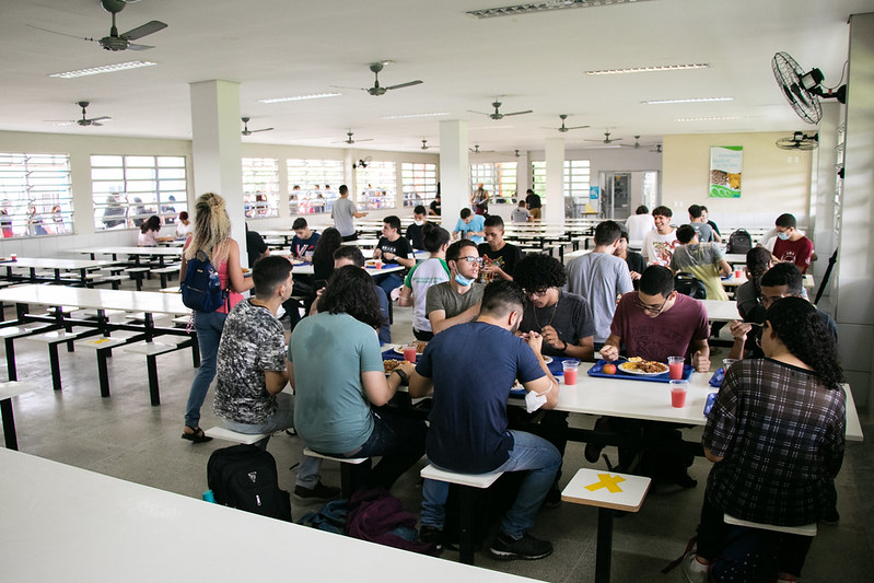Restaurantes Universitários da UFC em Fortaleza têm funcionamento modificado durante as férias acadêmicas, em julho (Foto: Ribamar Neto/UFC) Imagem: Foto interna do Restaurante Universitário do Campus do Pici da UFC, com mesa à direita ocupada por nove estudantes, uma aluna de blusa rosa e mochila preta caminhando no corredor e carregando uma bandeja, e mais três mesas ao fundo do refeitório, à direita, com alunos almoçando.