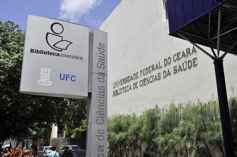 As bibliotecas universitárias também oferecerão horários especiais no período de férias (Foto: Guilherme Braga/UFC) Imagem:Fachada da Biblioteca de Ciências da Saúde da Faculdade Medicina da UFC, com placa na cor prata ao lado esquerdo da foto, identificada como “Biblioteca universitária UFC”, e plantas ornamentais do lado direito da foto.