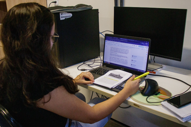 Imagem: Fotografia de uma pessoa sentada diante de uma mesa de trabalho em um escritório. Vista de costas e de perfil, ela utiliza um notebook enquanto consulta um documento impresso aberto ao lado do computador, segurando um marcador de texto amarelo. Na tela do notebook, há um editor de texto com um documento em elaboração. Sobre a mesa também estão um mouse, um monitor desligado, um computador de mesa, um smartphone e um caderno. O ambiente é iluminado por luz artificial e sugere uma atividade de revisão ou produção de documentos.