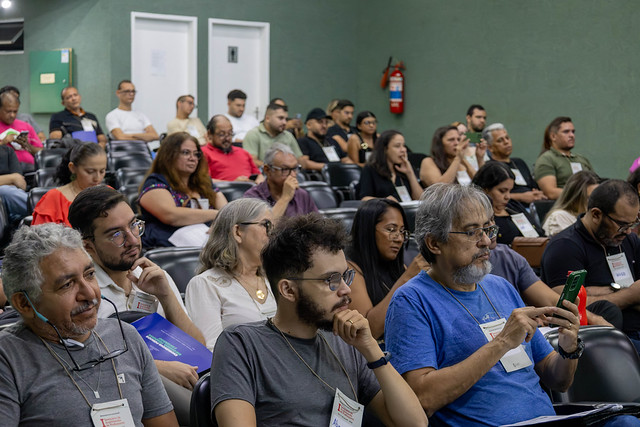 Imagem: Pessoas sentadas no Auditório Reitor Ícaro de Sousa Moreira acompanham a programação do Seminário de Acolhimento de Professores do Programa Mais Ciência na Escola – UFC. Os participantes observam a apresentação, enquanto alguns fazem anotações e utilizam aparelhos celulares