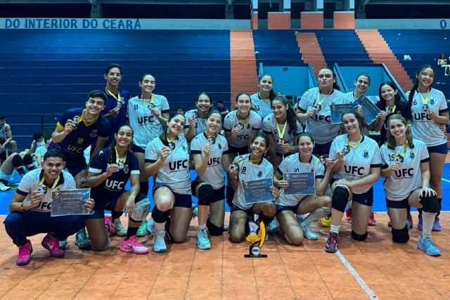 O pódio foi decidido nos jogos realizados no último domingo (26), em Aracati (Foto: Jefferson Gomes/Secretaria de Esportes UFC) Imagem: Trata-se de uma foto das atletas e da equipe técnica da seleção feminina de vôlei da UFC reunidas em quadra, exibindo medalhas, troféu e certificados conquistados. O grupo posa sorridente em um ginásio, celebrando o resultado da competição.